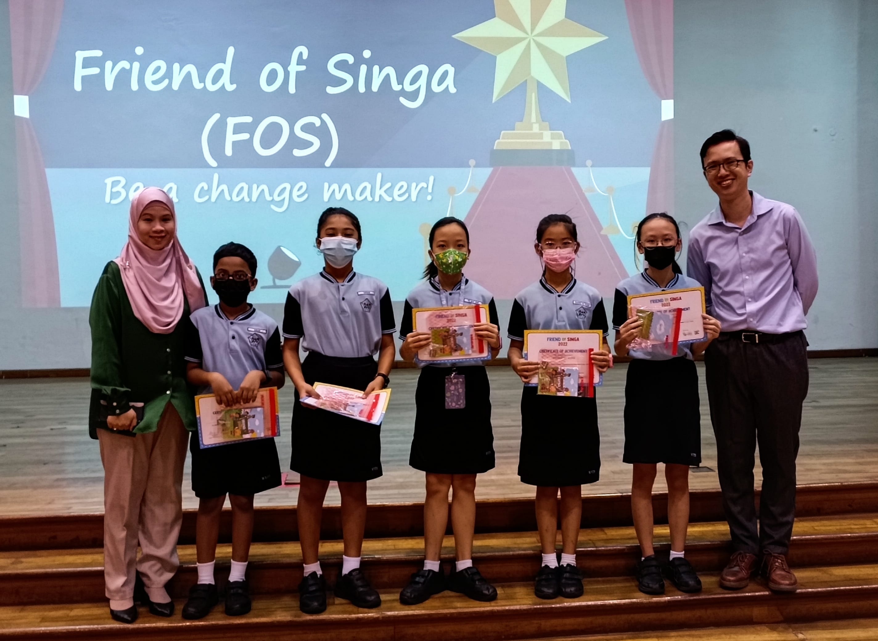Five masked students hold certificates beside two adults in front of a Friend of Singa FOS Be a change maker slide.